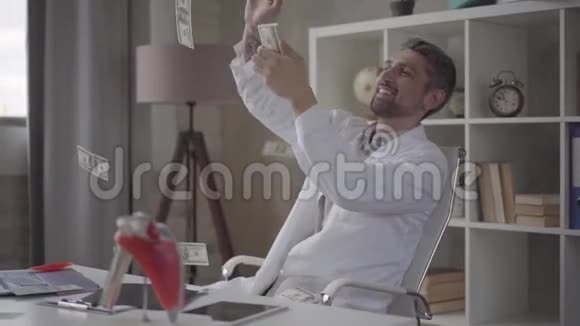 英俊的快乐的成功的满意的留胡子的医生把坐在椅子上的撒在工作桌上视频的预览图
