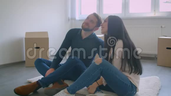 年轻的白种人夫妇正坐在靠近窗户的新房子的地板上还有梦想着新家生活的纸盒视频的预览图