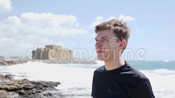 年轻的帅哥在跑步锻炼前戴上耳机站在海滩上微笑背景上的城堡慢动作视频的预览图