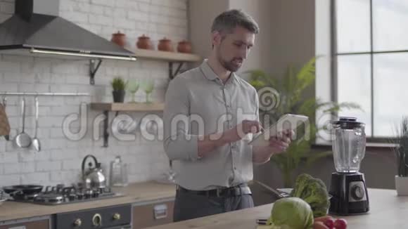 英俊帅气的男人穿着衬衫做饭在家厨房里检查平板电脑上的食谱概念视频的预览图