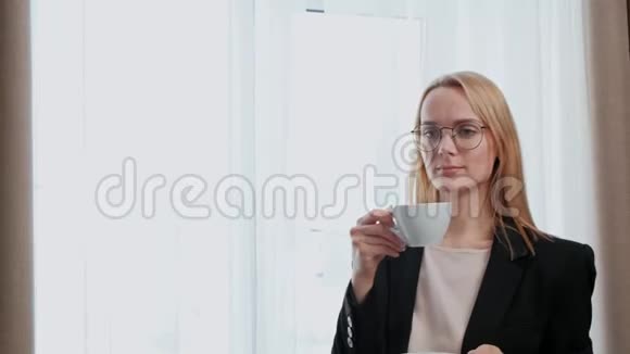 年轻迷人的中年金发女人穿着黑色商务套装戴着酒店房间的眼镜站在窗边视频的预览图