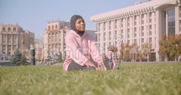 城市中戴着头巾坐在草地上的年轻美籍黑人穆斯林女孩的特写照片视频的预览图