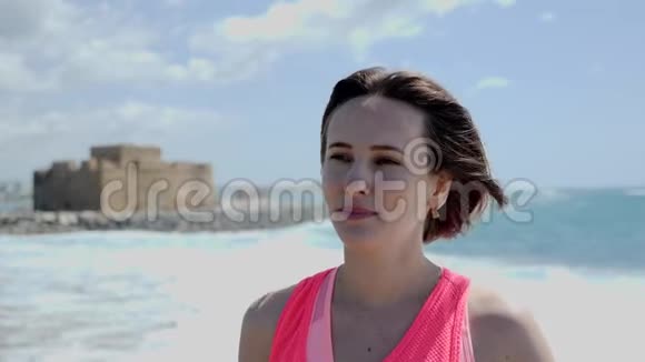 年轻漂亮的女人在海滩上训练前戴上耳机强浪正在撞击海岸慢动作视频的预览图