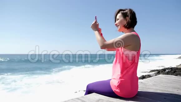 年轻美丽的白种人女人坐在岩石海滩上拿着电话拍着海浪拍打岩石的照片视频的预览图