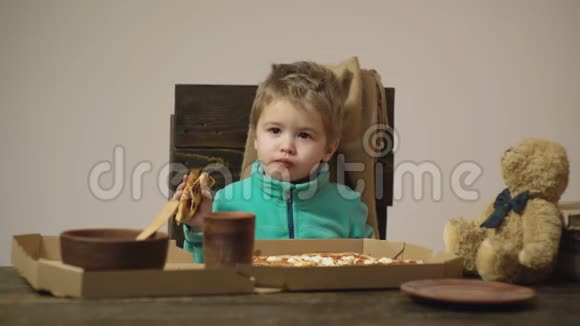 可爱的小白种人男孩在木桌上吃披萨上面有披萨盒勺子盘子和熊视频的预览图