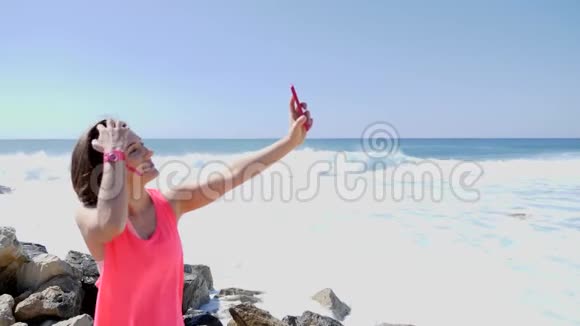 年轻美丽的白种人女孩带着电话做自拍照片而站在岩石海滩上抚摸她的头发微笑慢节奏视频的预览图