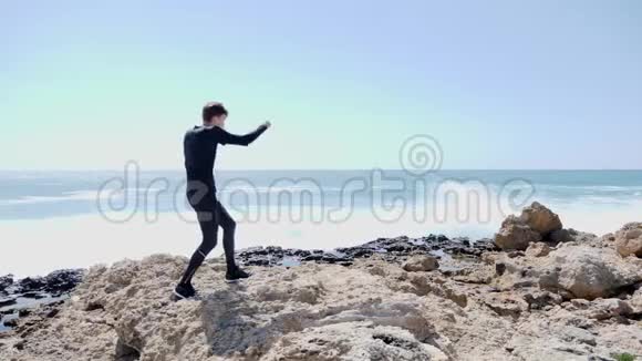 年轻苗条的男子拳击空气在岩石海滩强浪正在撞击海岸风和日丽的一天慢动作视频的预览图