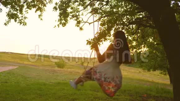 美丽的女孩穿着裙子在公园里的秋千上飞翔年轻女孩在橡木树枝上的绳索秋千上摆动少女少女少女视频的预览图