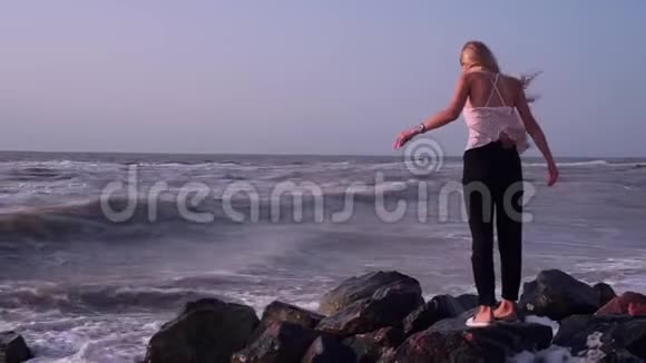 穿着T恤的金发女郎站在海里的岩石上被海浪喷洒视频的预览图