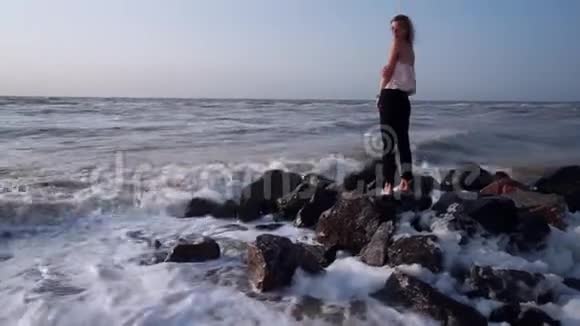 穿T恤的漂亮女孩站在海中的岩石上在海浪中飞溅着视频的预览图