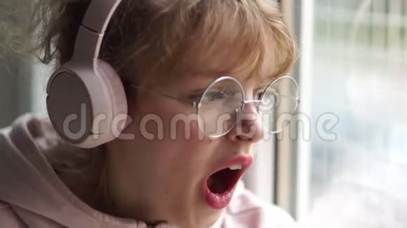 穿着粉红色耳机和眼镜的少女无聊地坐在家靠近窗户女孩打哈欠捂住嘴视频的预览图