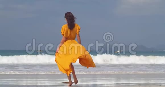 优雅的年轻女士穿着长长的黄色连衣裙在潮湿的沙滩上慢慢地走着在海边享受美好的一天视频的预览图