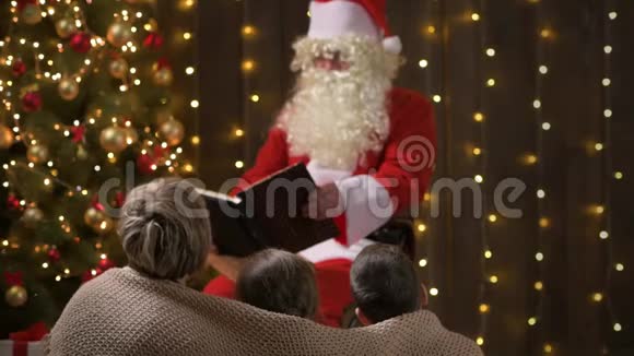圣诞老人给家人看书母亲和孩子们坐在室内靠近装饰好的圣诞树灯光和倾听圣诞快乐视频的预览图
