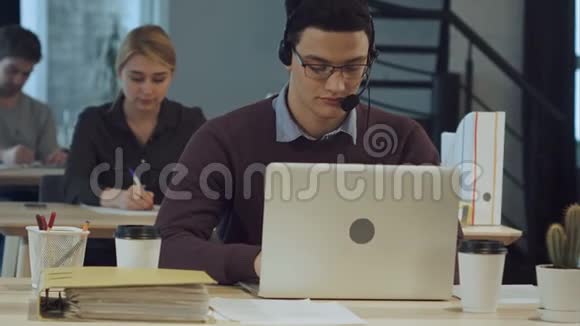 在电话中心工作的快乐的年轻工人带着耳机微笑在笔记本电脑上工作视频的预览图