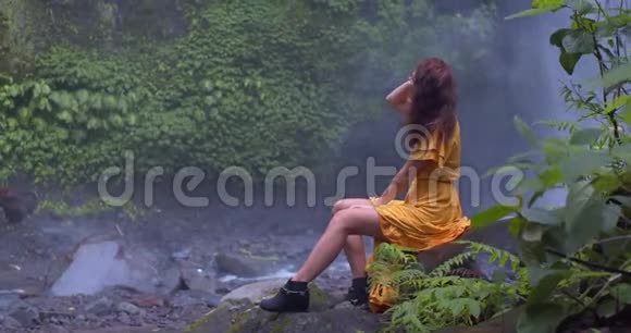 可爱的女孩穿着黄色的裙子坐在瀑布下湿漉漉的岩石风摇曳着她的头发她一只手把它固定下来视频的预览图