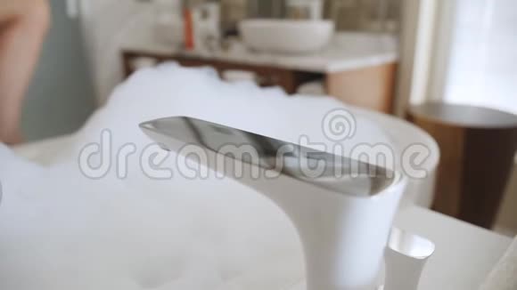 一个女孩坐在一个充满泡沫的浴室里前景中鹤的特写镜头和坐在浴缸里的女孩模糊了视频的预览图