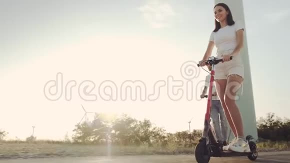 日落时分一个男孩和一个女孩在风力发电机的背景下骑着电动滑板车视频的预览图