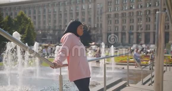 站在城市喷泉旁边的年轻漂亮的戴头巾的非裔美国穆斯林女孩的特写照片视频的预览图
