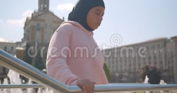 在城市室外的喷泉旁伸展着迷人的戴头巾的非裔美国穆斯林女孩的特写肖像视频的预览图