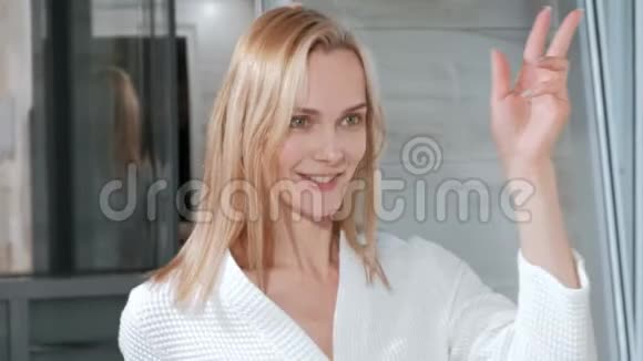 漂亮的中年金发女人穿着白色浴袍站在镜子旁边的浴室里有趣的舞蹈和微笑视频的预览图