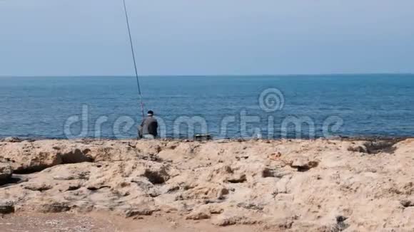 穿着深色毛衣戴着钓鱼竿的帽子头发灰白的老渔夫坐在海边钓鱼视频的预览图