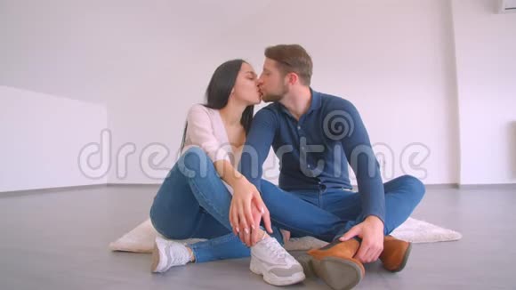 在白墙背景下一对正在进行的白种人夫妇坐在新的空平拥抱和亲吻的地板上快乐着视频的预览图