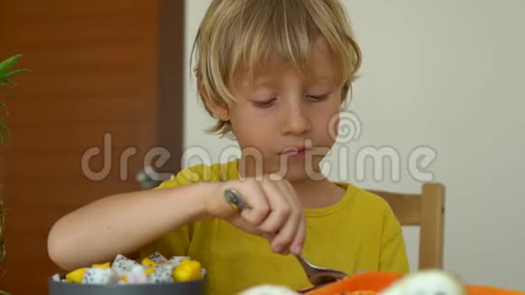 一个穿黄色衬衫的小男孩吃一种激情水果或马拉库贾许多放在桌子上的热带水果围绕着他视频的预览图