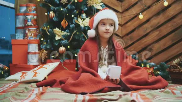 4k视频可爱的微笑女孩坐在舒适的家里喝热巧克力在圣诞树上的木制背景视频的预览图
