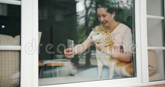 美丽的女孩和狗在窗台的咖啡馆里用智能手机自拍视频的预览图