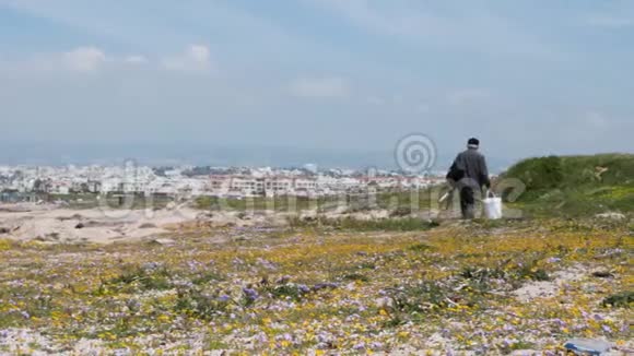 穿深色衣服的老渔夫戴着鱼竿和水桶沿着海滨散步老年男性穿过岩石海滩视频的预览图