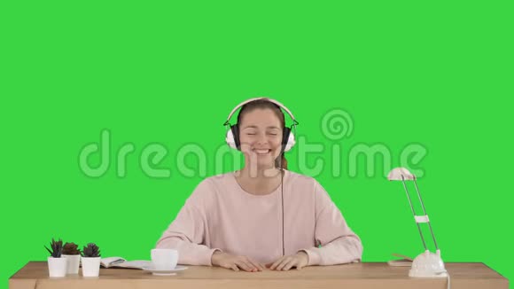 漂亮的女士穿着粉红色套头衫听着耳机里的音乐微笑着在绿色屏幕上拍摄Chroma键视频的预览图