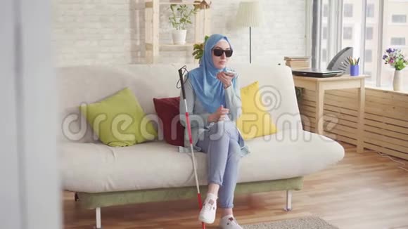 漂亮的穆斯林盲女在家戴着头巾的拐杖坐在沙发上享受智能手机视频的预览图