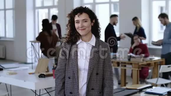 在现代办公室里一位留着卷发对着镜头微笑的快乐积极向上的年轻白人商业女性的中型肖像画视频的预览图