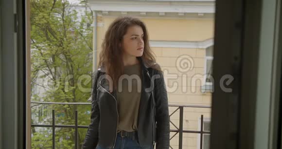 穿着皮夹克的漂亮黑发女孩站在阳台上看到绿色的城市景色微笑着进入镜头高兴和快乐视频的预览图