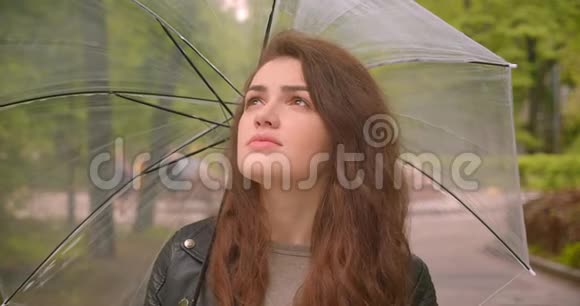 在绿色城市公园里白种人深色头发的女性在伞下用平静的微笑观看镜头视频的预览图