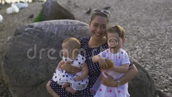 年轻的母亲带着她的小女儿微笑着在河边看着相机穿着点缀的裙子家庭价值观视频的预览图