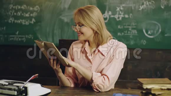 穿着粉红色衬衫微笑的年轻女子正在绿色黑板附近读一本书上面写着公式年轻漂亮视频的预览图