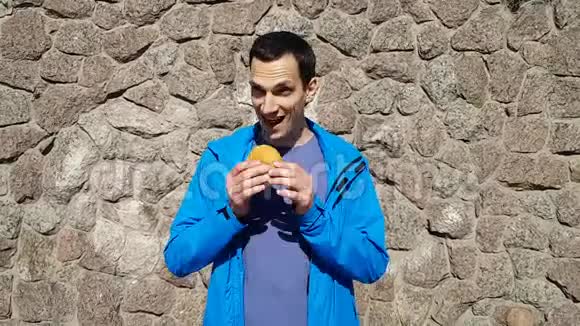 有趣的开朗的黑发男人在街上靠石头墙吃一个小汉堡包快速小吃视频的预览图