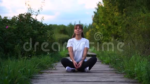 年轻漂亮的女孩坐在木桥上摆出莲花的姿势用慢动作微笑着向太阳问好视频的预览图