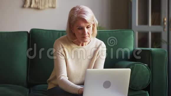 坐在沙发上的一位严肃的年长的成熟女人在电脑上打字视频的预览图