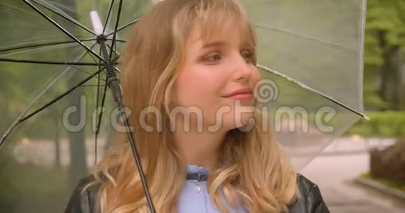 漂亮的白种人金发学生带着雨伞害羞地微笑着进入绿色城市公园的镜头视频的预览图