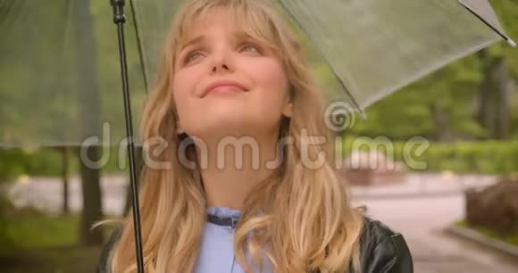 在绿色城市公园里漂亮的白种人金发学生兴高采烈地拿着雨伞对着镜头视频的预览图