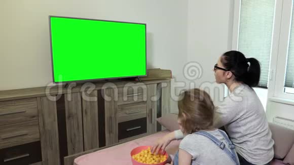 女儿和母亲从碗里吃薯片并在电视旁使用带有绿色屏幕的遥控器视频的预览图