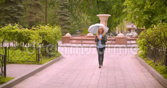 美丽的白种人金发学生带着雨伞走在绿色公园观察大自然的美丽微笑着进入镜头视频的预览图