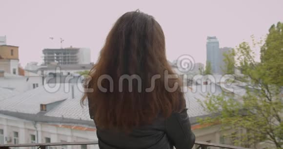 阳台上穿着皮夹克的黑发女孩坐在阳台上欣赏着都市的景色后镜头转向镜头兴高采烈地微笑着视频的预览图