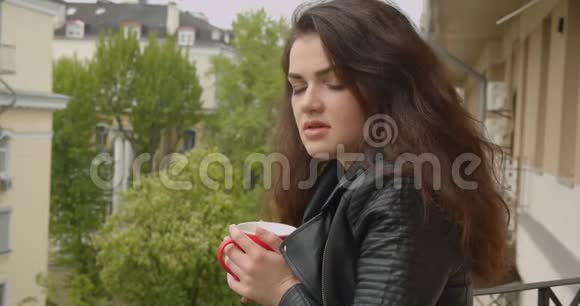 年轻的黑发女孩穿着皮夹克从阳台上欣赏壮观的绿色城市景色喝着热茶视频的预览图