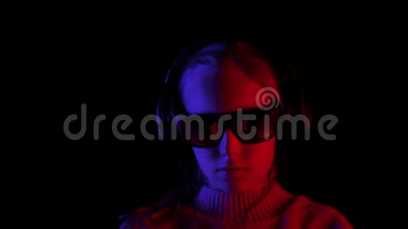 微笑的女孩听着耳机和黑色太阳镜的移动音乐在黑暗的工作室红色和蓝色的背光视频的预览图