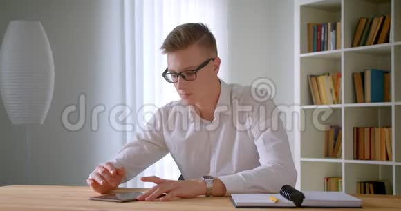 年轻英俊的白种人商人戴着眼镜使用平板电脑和在室内做笔记的特写照片视频的预览图