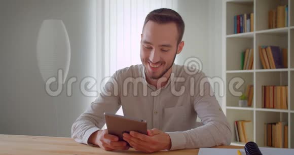 年轻英俊的白种人商人用平板电脑在室内愉快地看着摄像机的特写镜头视频的预览图