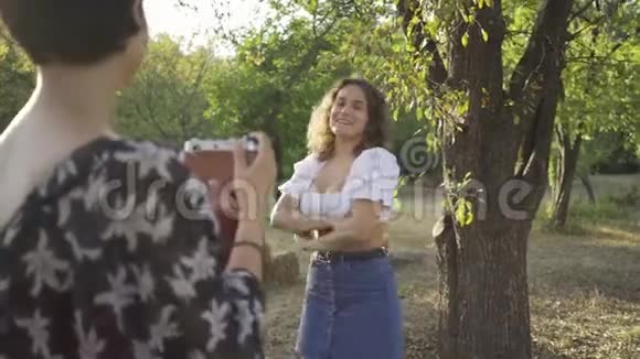 白人女性留着卷发在背景中摆姿势而她的朋友在前景中使用旧相机拍照视频的预览图